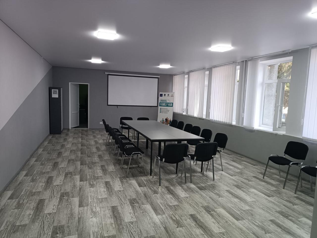 Світла, сучасна конференц-зала із довгим столом, чорними стільцями, проекційним екраном та банером Family Hub біля вікон із жалюзі.