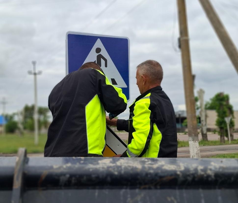 Двоє чоловіків у чорно-жовтих робочих куртках встановлюють дорожній знак Пішохідний перехід на узбіччі.