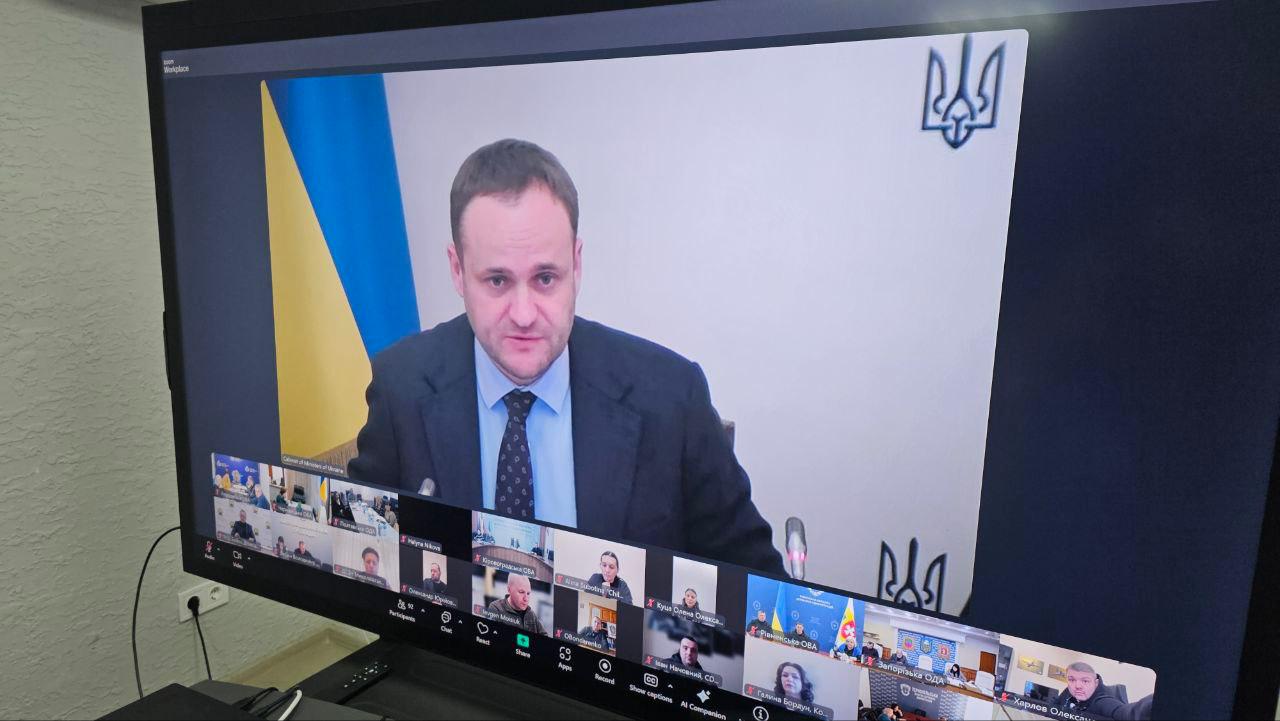 Монітор комп'ютера, на якому відображається вікно відеоконференції з головним доповідачем у центрі та іншими учасниками у нижній частині екрана. На фоні відео доповідача видно державний прапор України, а у верхньому куті монітора — малий герб України.