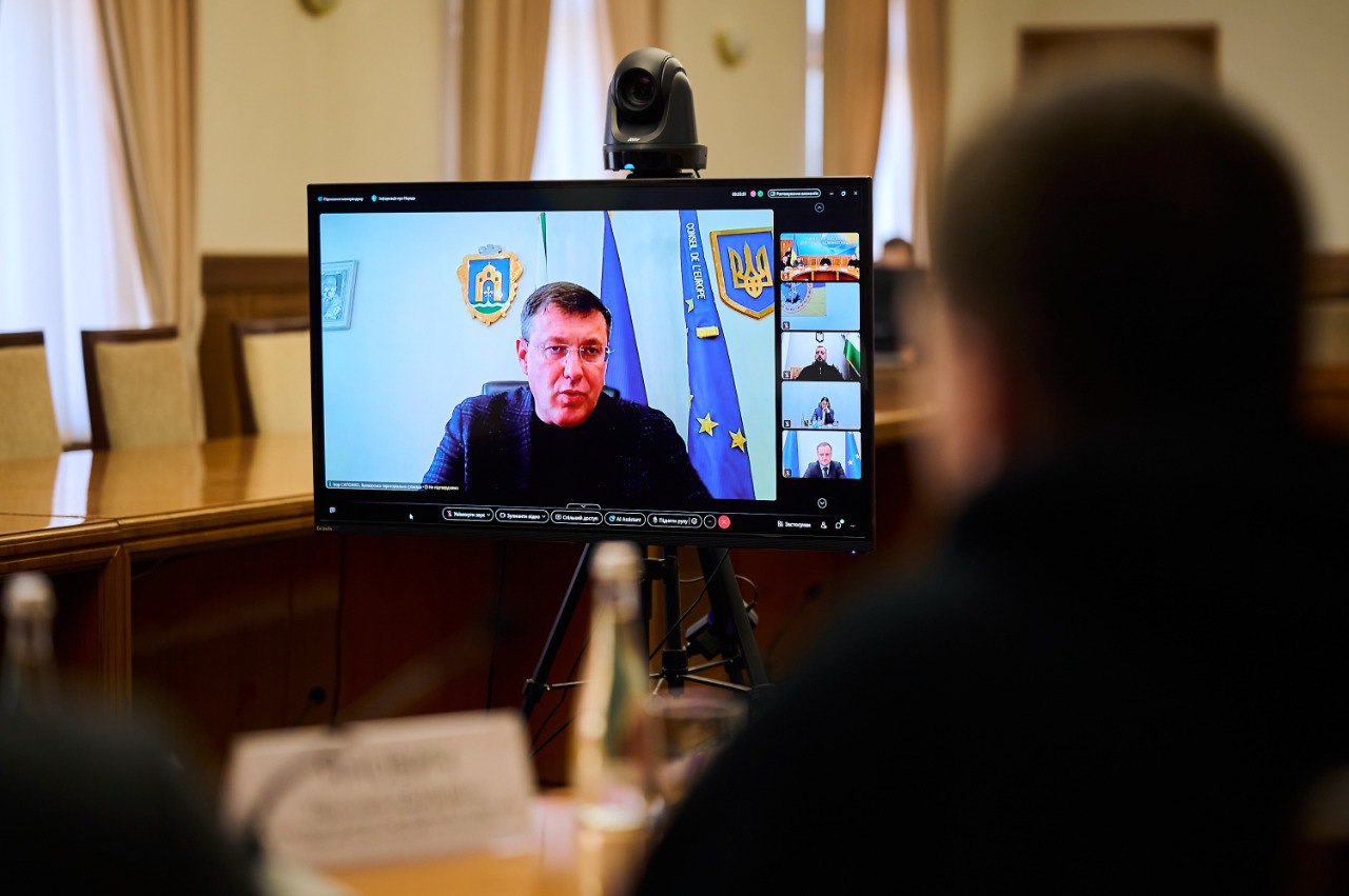 На екрані монітора під час відеоконференції видно чоловіка у діловому одязі. На задньому плані зображення з екрана — прапор України та герб.