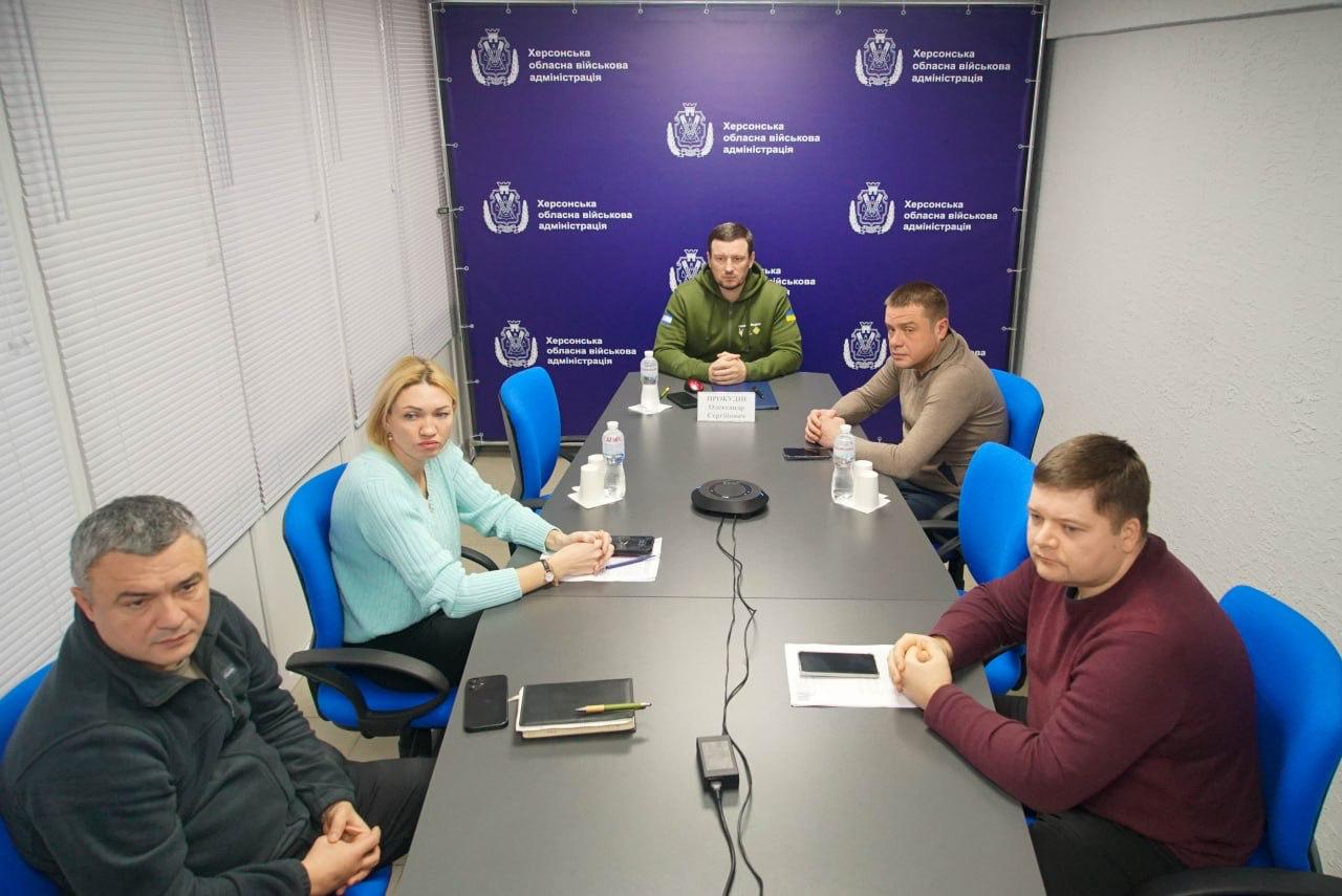 Група з п'яти осіб, четверо чоловіків і одна жінка, сидять за столом для нарад. На синьому тлі позаду них розміщені логотипи та написи: Херсонська обласна військова адміністрація.