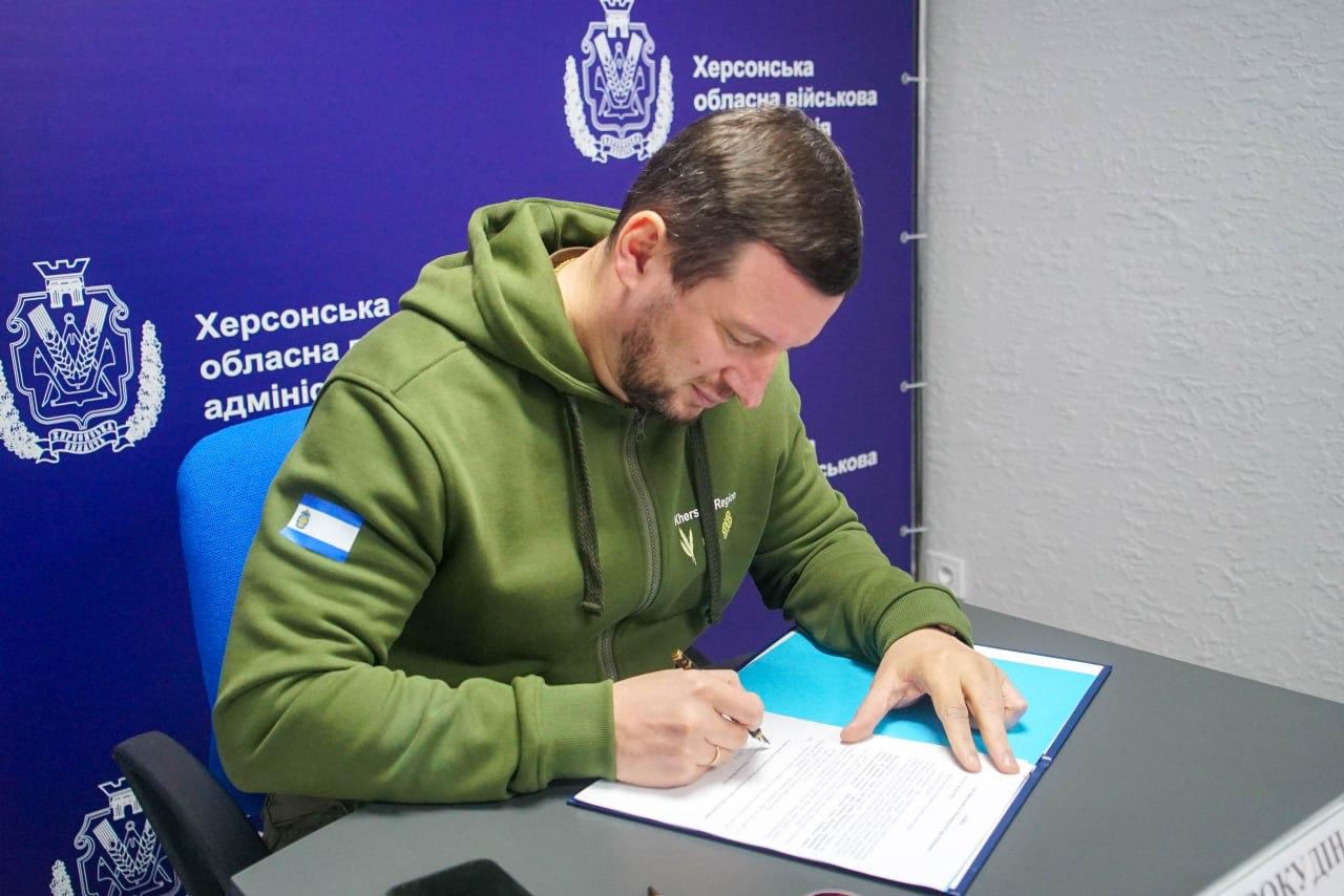 Чоловік у зеленому худі сидить за столом і підписує документ у синій папці. На синьому тлі позаду нього видно логотипи та написи: Херсонська обласна військова адміністрація.