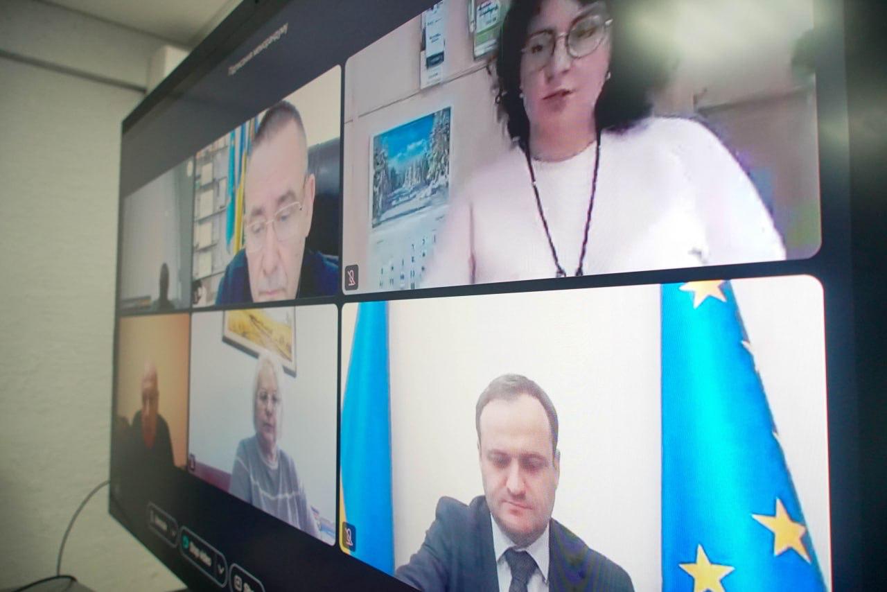 Відеоконференція за участі шести осіб, які з'являються у вікнах на екрані монітора. Один чоловік сидить на тлі українського та європейського прапорів.