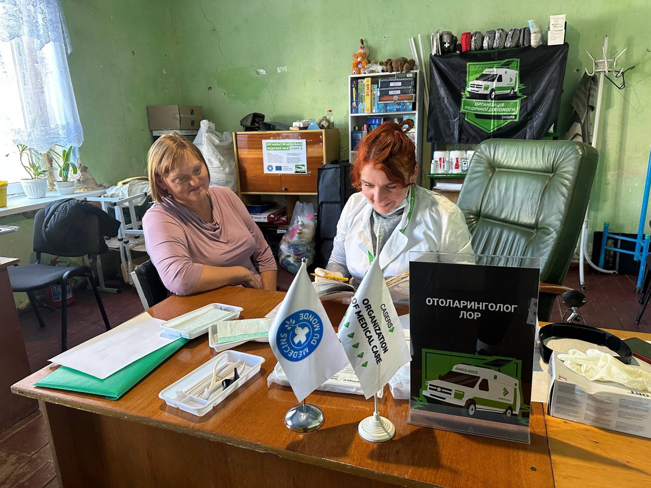 Лікар-отоларинголог за столом спілкується з пацієнткою, перед ними стоять прапорці: CASERS та MEDECA, а також табличка ОТОЛАРИНГОЛОГ ЛОР.