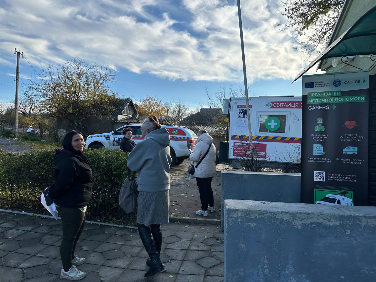 Кілька жінок стоять на вулиці біля мобільного медичного пункту СВІТАЛИЦЯ та автомобіля LEGACY OF WAR, що надає медичну допомогу.