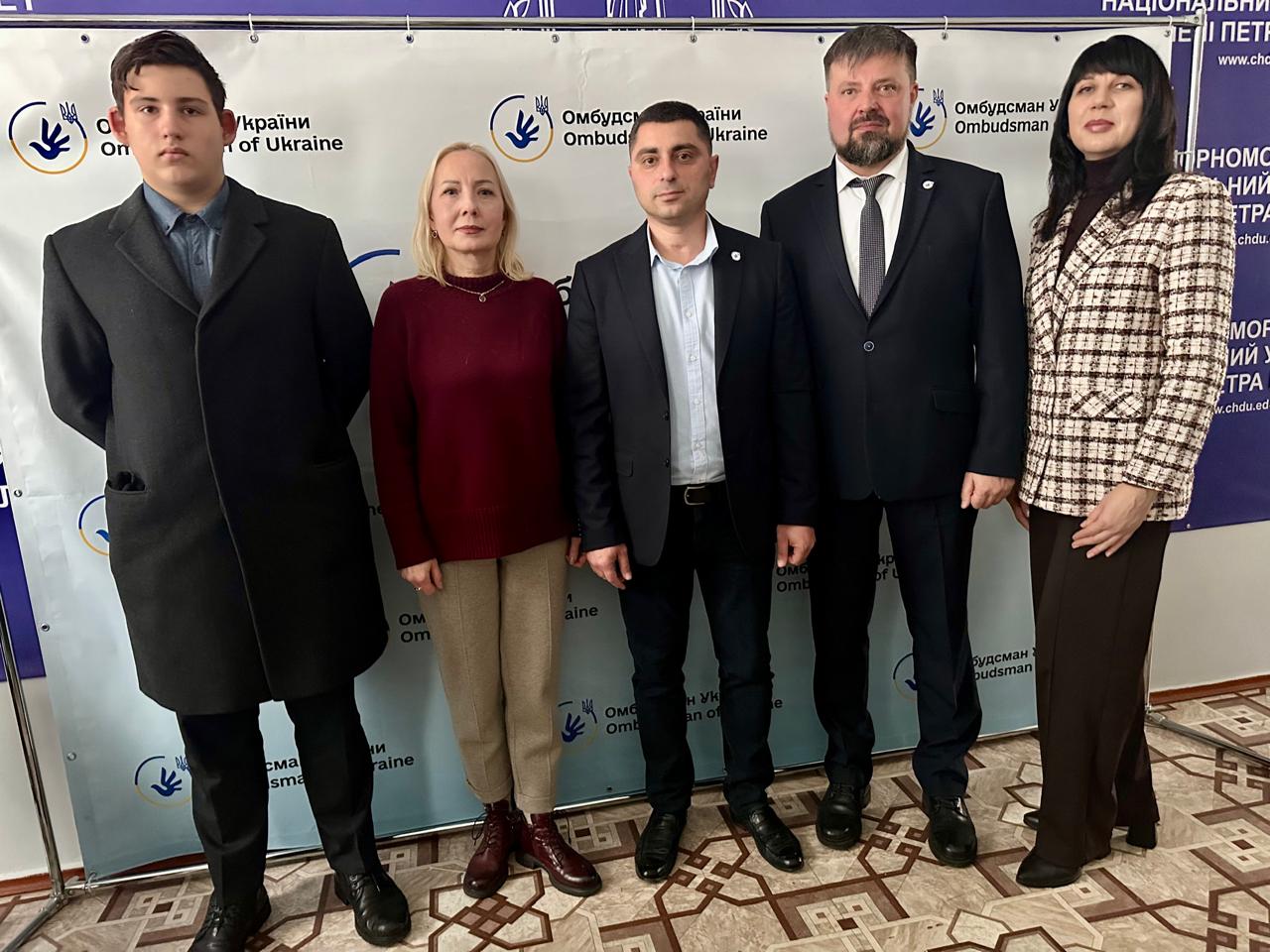 П'ятеро людей — троє чоловіків і дві жінки — стоять разом перед банером, на якому видно логотип та написи: Омбудсман України та Ombudsman of Ukraine.