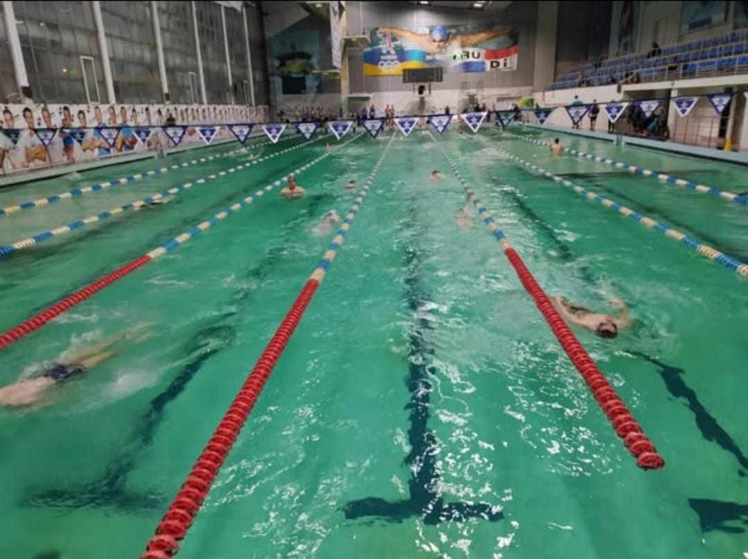 Довгий критий басейн із кількома доріжками, де плавають спортсмени; басейн огороджений червоними, білими та синіми буями.