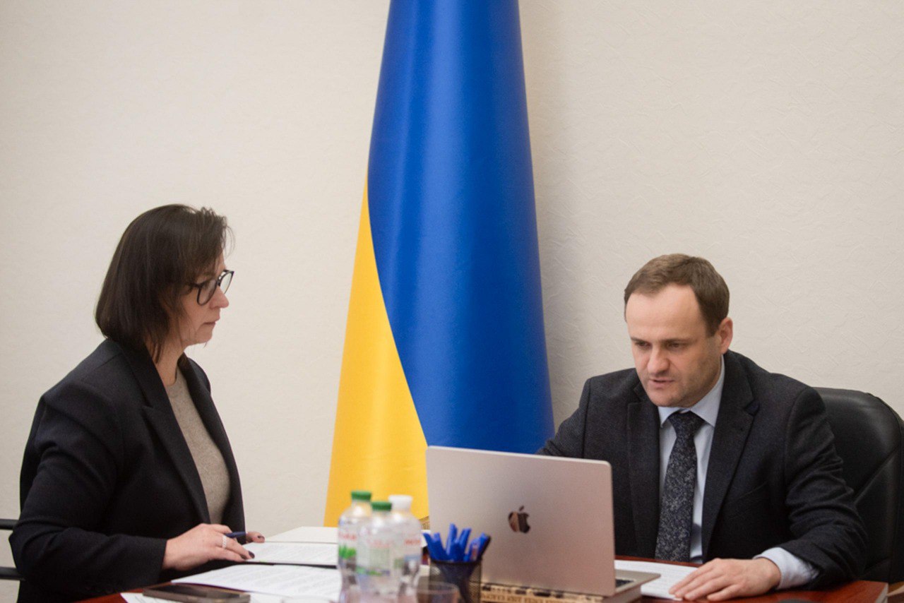 Двоє людей, чоловік у костюмі та жінка в жакеті й окулярах, сидять за столом, працюючи з ноутбуком та документами. На задньому плані розташований прапор України.