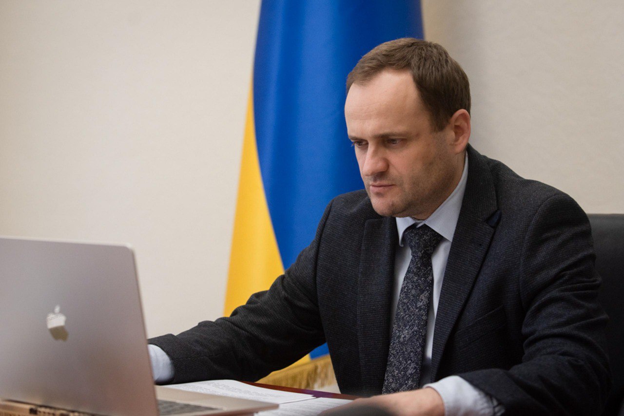 Чоловік у діловому костюмі сидить за столом, дивлячись на ноутбук. На задньому плані видно прапор України.