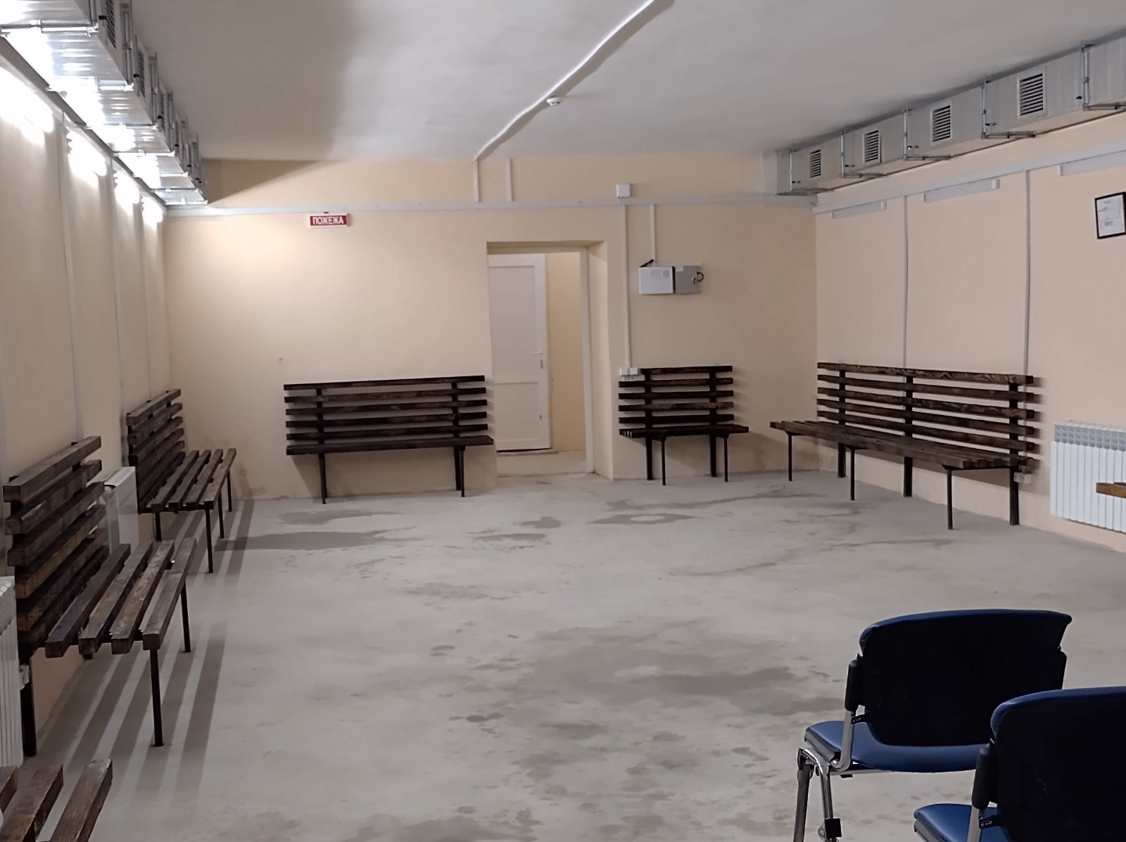 Просторе, світле приміщення очікування зі світло-бежевими стінами та цементною підлогою. Уздовж стін розташовані дерев'яні лавки з темними рейками. Над дверима у центрі приміщення висить маленька червона табличка з написом: ПОЖЕЖА. Під стелею видно металеві вентиляційні отвори та ряди люмінесцентних світильників. На передньому плані видно частину синіх офісних стільців.