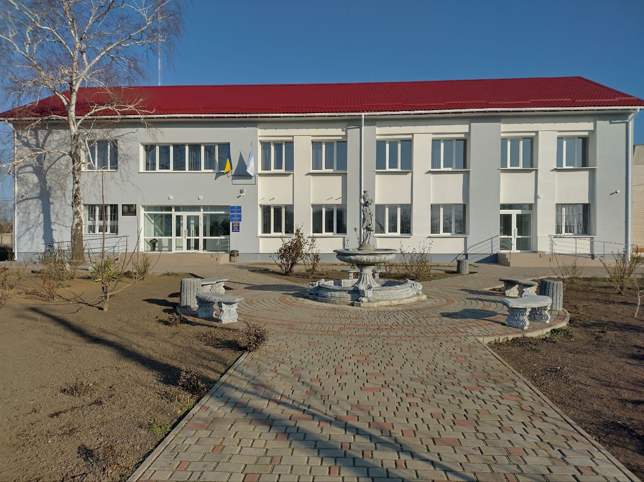 Двоповерхова адміністративна будівля зі світло-сірими стінами та яскраво-червоним дахом у сонячний день. Перед входом розташована вимощена цеглою площадка з декоративним фонтаном посередині та кам'яними лавками. На фасаді будівлі видно прапори України та, ймовірно, інший прапор чи емблема. Навколо фонтану знаходиться невеликий сквер з голими деревами та кущами.