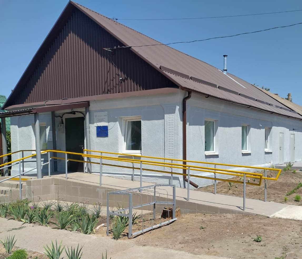 Фасад одноповерхової адміністративної будівлі зі світло-сірими стінами та коричневим профнастиловим дахом. Зліва облаштовано пандус із жовтими поручнями для забезпечення доступності. На стіні біля входу розташована невелика синя табличка, а поруч із будівлею видно свіжі насадження та голі гілки.