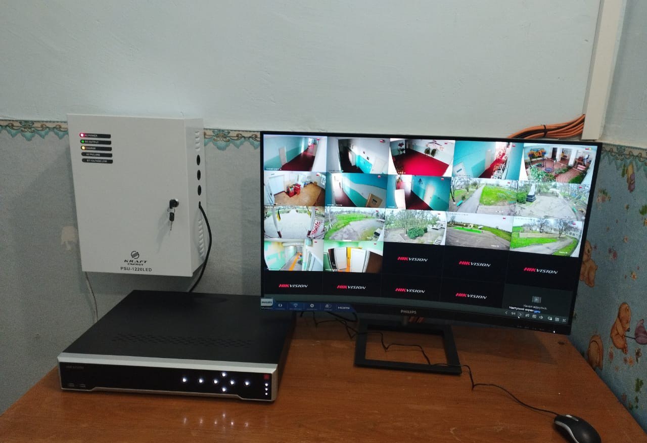 Система відеоспостереження, встановлена на столі, що складається з чорного відеореєстратора Hikvision, монітора Philips, на якому відображається сітка з 16 зображеннями з камер, та білого настінного блоку живлення KRAFT PFU-120LED. Стіни мають світло-блакитний колір, а стіл виготовлено з дерева.