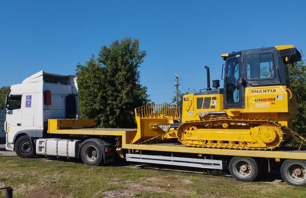 Жовтий гусеничний бульдозер SHANTUI моделі DH13-B2 XL завантажено на причіп-платформу, яку тягне біла вантажівка-тягач, і він транспортується по ґрунтовій ділянці. На задньому плані видно зелені дерева та блакитне небо.