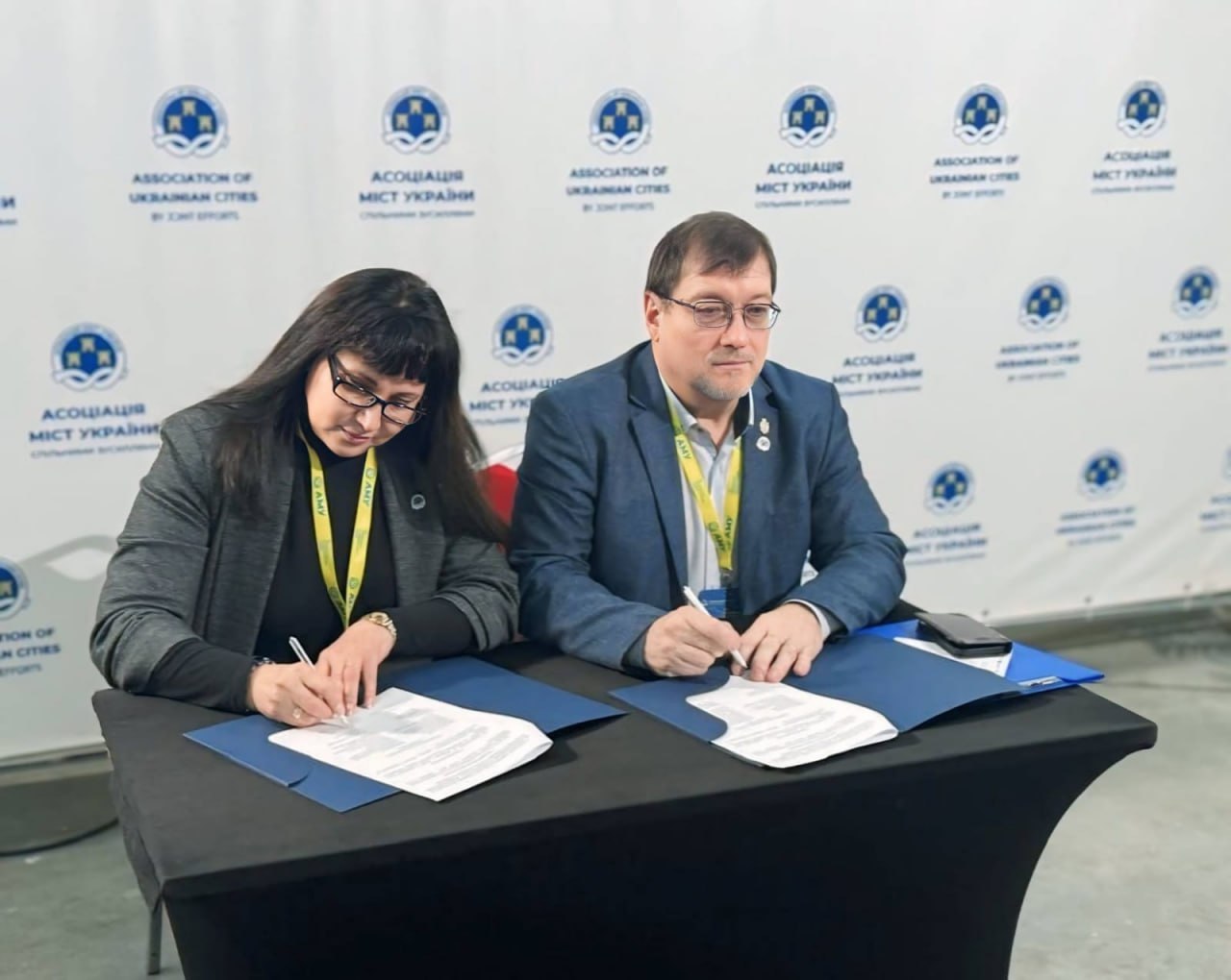 Жінка та чоловік в діловому одязі сидять за столом, підписуючи документи на фоні банера з логотипом: Асоціація міст України (Association of Ukrainian Cities).