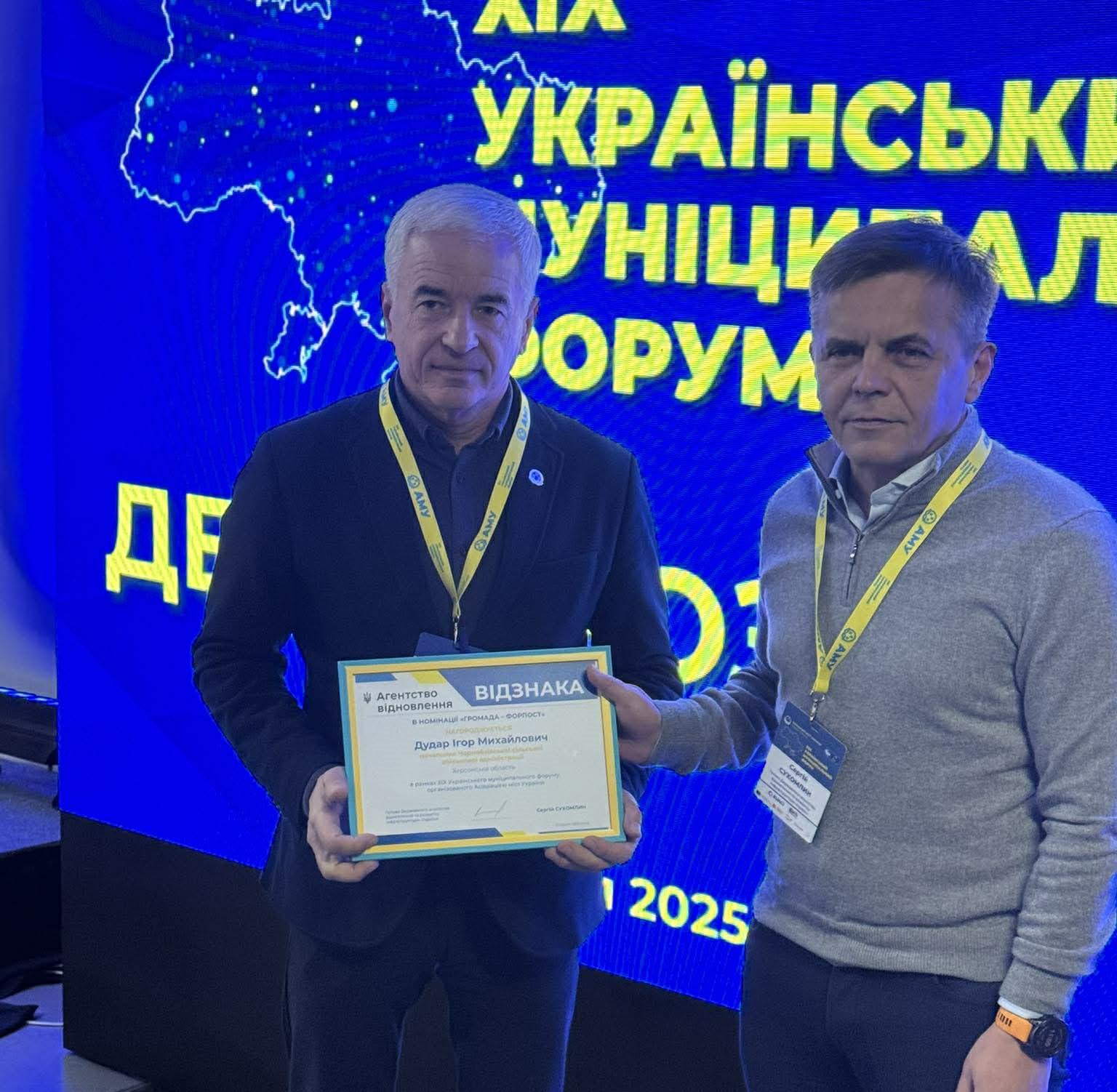 На світлині двоє чоловіків стоять на тлі синього екрана з логотипом Асоціації міст України. Чоловік у центрі, одягнений у чорне, тримає грамоту-відзнаку, яку йому вручає інший чоловік у сірому светрі.