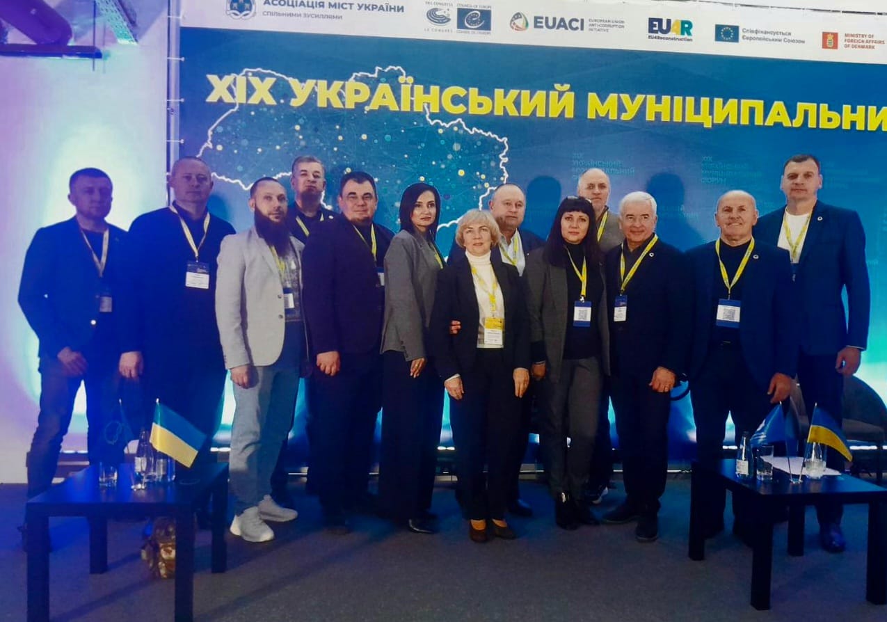 Група з приблизно дванадцяти людей (чоловіки та жінки) стоять разом і дивляться в камеру на тлі великого синьо-жовтого банера.