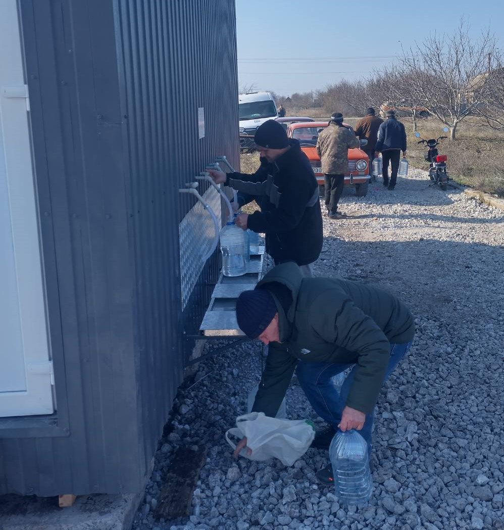 Двоє чоловіків набирають питну воду у великі пластикові бутлі з системи зовнішніх кранів, встановленої на стіні темно-сірого модульного приміщення. На задньому плані видно ще кількох людей, які стоять поруч із припаркованими автомобілями (зокрема, помаранчевою старою моделлю) та мотоциклом на гравійній ділянці.