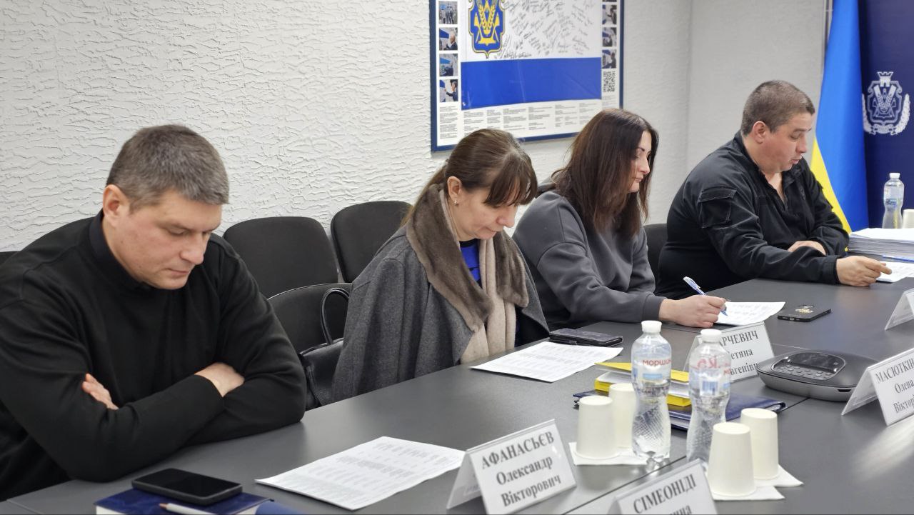 На світлині видно чотирьох учасників наради, які сидять за столом, зосереджено працюючи з документами. На задньому плані розташовані плакати з символікою України та Херсонської області.