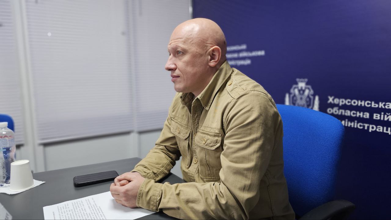Портретний знімок чоловіка середнього віку з лисою головою, який сидить за столом у профіль, дивлячись ліворуч. Він одягнений у світло-коричневу сорочку мілітарі стилю з кишенями. Перед ним на столі лежать документи та мобільний телефон. На задньому плані видно синій банер з написом: Херсонська обласна військова адміністрація.