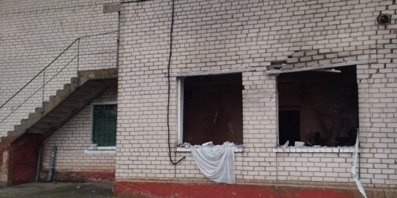 Фасад сильно пошкодженої (ймовірно, внаслідок обстрілу) Пункту незламності з побіленої цегли, де видно два великі прямокутні отвори від зруйнованих вікон. Зліва на задньому плані розташовані зовнішні бетонні сходи. Віконні отвори не мають скла, видно уламки та внутрішні приміщення. Стіни мають сіро-білий колір, а нижня частина фасаду пофарбована у червоний.