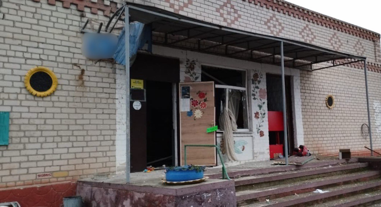 Фасад сильно пошкодженого обстрілом Пункту незламності з бежевої цегли. Вхідна зона, віконні отвори та двері зруйновані: вікна розбиті, видно уламки та внутрішні приміщення. Над входом є навіс, а стіна навколо вікна прикрашена декоративними квітковими візерунками. Зліва видно частину прикраси у вигляді жовтої шини-соняшника.