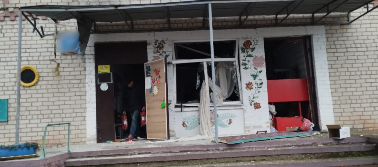 Фасад сильно пошкодженого обстрілом Пункту незламності з бежевої цегли. Віконні отвори та вхідні двері зруйновані: вікна розбиті, внутрішні приміщення видно через отвори, а поруч зі вхідними дверима лежать уламки. Стіна біля вікна частково пофарбована декоративними квітковими візерунками. Біля входу стоїть людина, яка тримає вогнегасник.