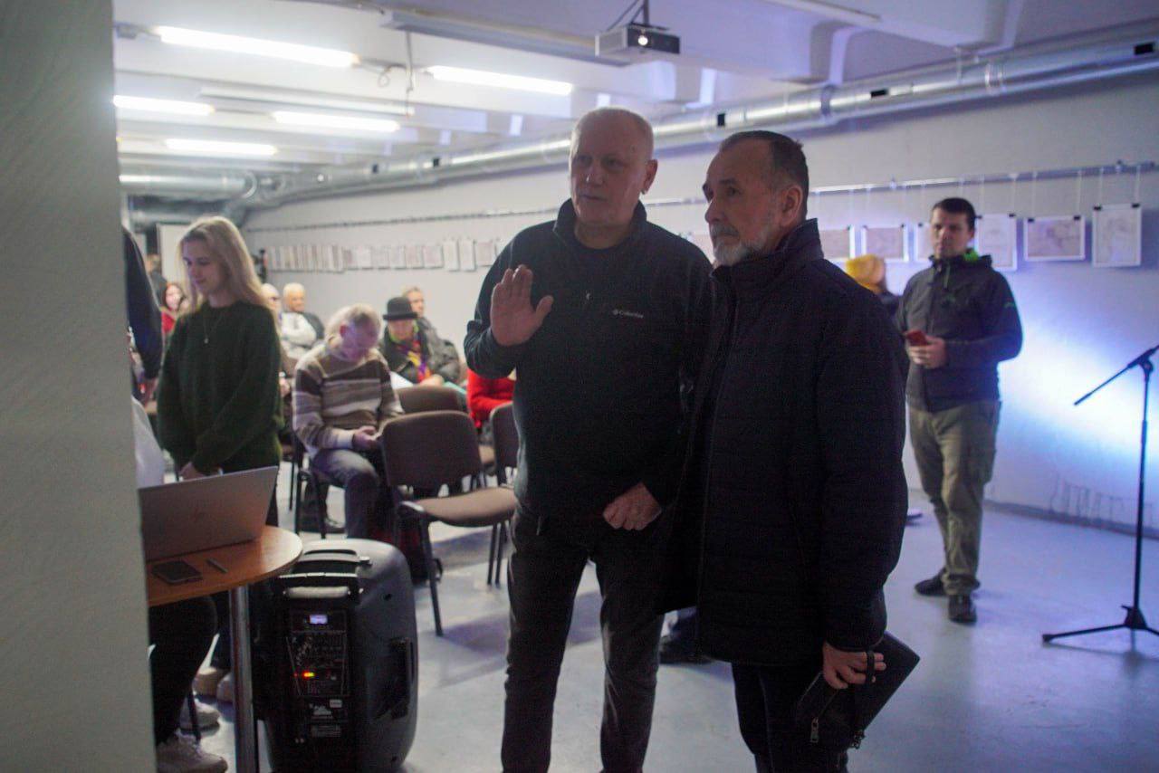 Двоє чоловіків стоять у приміщенні під час заходу: старший чоловік у чорній флісовій кофті з короткою стрижкою піднімає праву руку, а поруч стоїть чоловік із бородою в чорній куртці, який тримає невелику сумку. На задньому плані видно інших учасників, що сидять або стоять, та проекційне обладнання.