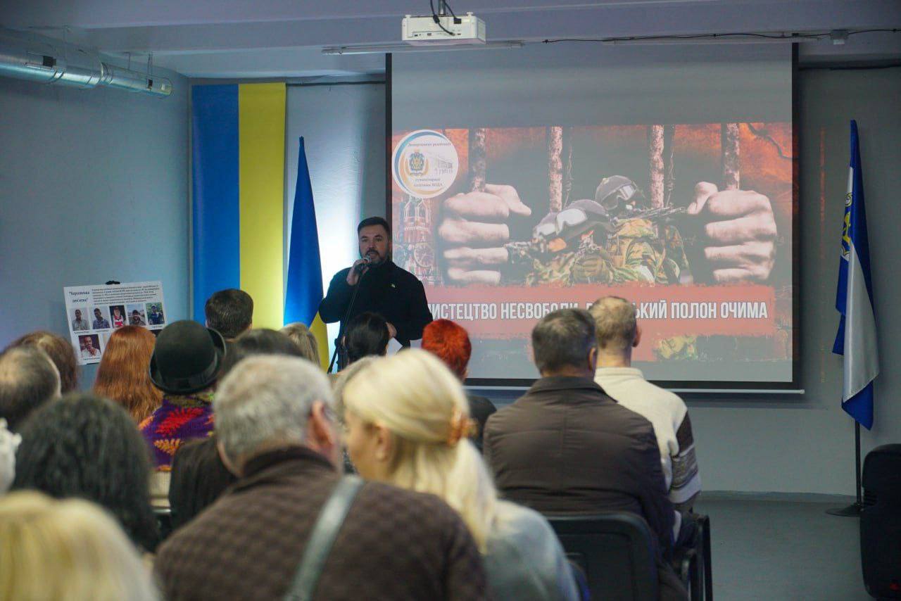 Чоловік із бородою стоїть перед проєкційним екраном і звертається до аудиторії, тримаючи мікрофон. На екрані велике зображення військових у касках за ґратами та напис, частина якого: РОСІЙСЬКИЙ ПОЛОН ОЧИМА ЦИВІЛЬНОГО. На задньому плані видно прапори України.