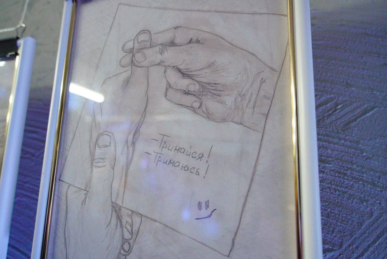 Графічний малюнок олівцем у рамці, що зображує руки, які тримають аркуш паперу з написаним від руки українським текстом: -Тримайся! -Тримаюсь! та простим смайликом знизу.