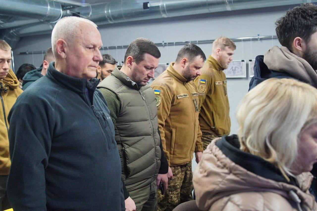 Група людей у цивільному та військовій формі, переважно чоловіки, стоять, схиливши голови, ймовірно, під час хвилини мовчання. Двоє чоловіків у центрі вдягнені у військові куртки кольору хакі та оливи.