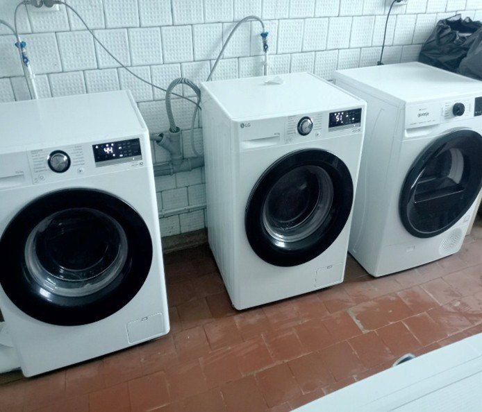 Три сучасні білі пральні машини стоять у ряду біля стіни, обкладеної білою плиткою. На підлозі - коричнева плитка. Одна з машин - сушильна.