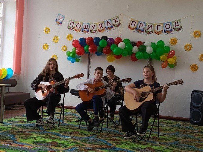 Четверо музикантів — троє дітей та підлітків з акустичними гітарами та одна доросла жінка за синтезатором — виступають у кімнаті, прикрашеній повітряними кульками та банером У ПОШУКАХ ПРИГОД.