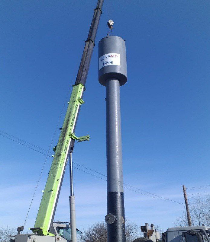 Кран піднімає або встановлює високу сіру циліндричну водонапірну башту на довгому стовпі. На циліндрі є написи: USAID та HOPE. Небо яскраво-блакитне.