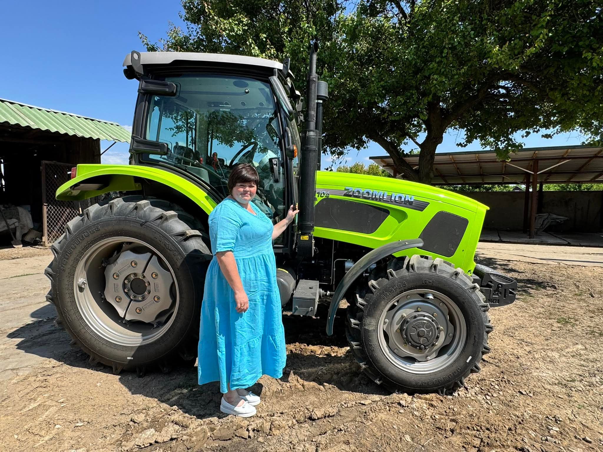 Жінка у блакитній сукні стоїть поруч із яскраво-зеленим трактором під деревом у сонячний день.