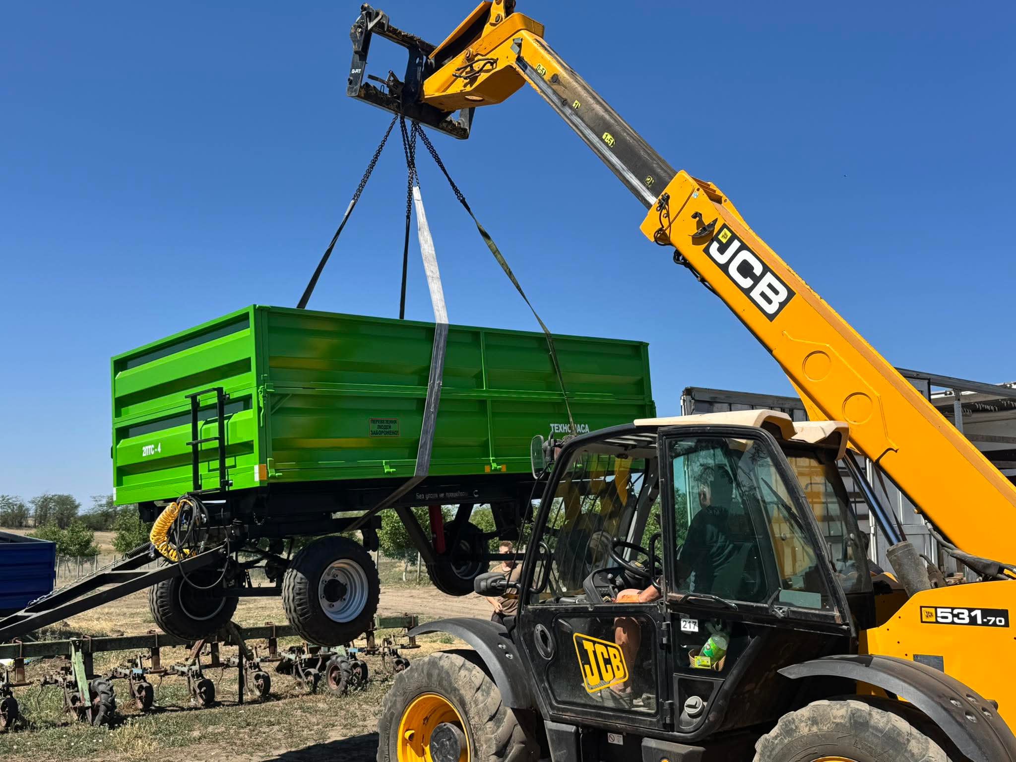 Жовто-чорний телескопічний навантажувач JCB підіймає великий зелений причіп для сільськогосподарської техніки на тлі блакитного неба.