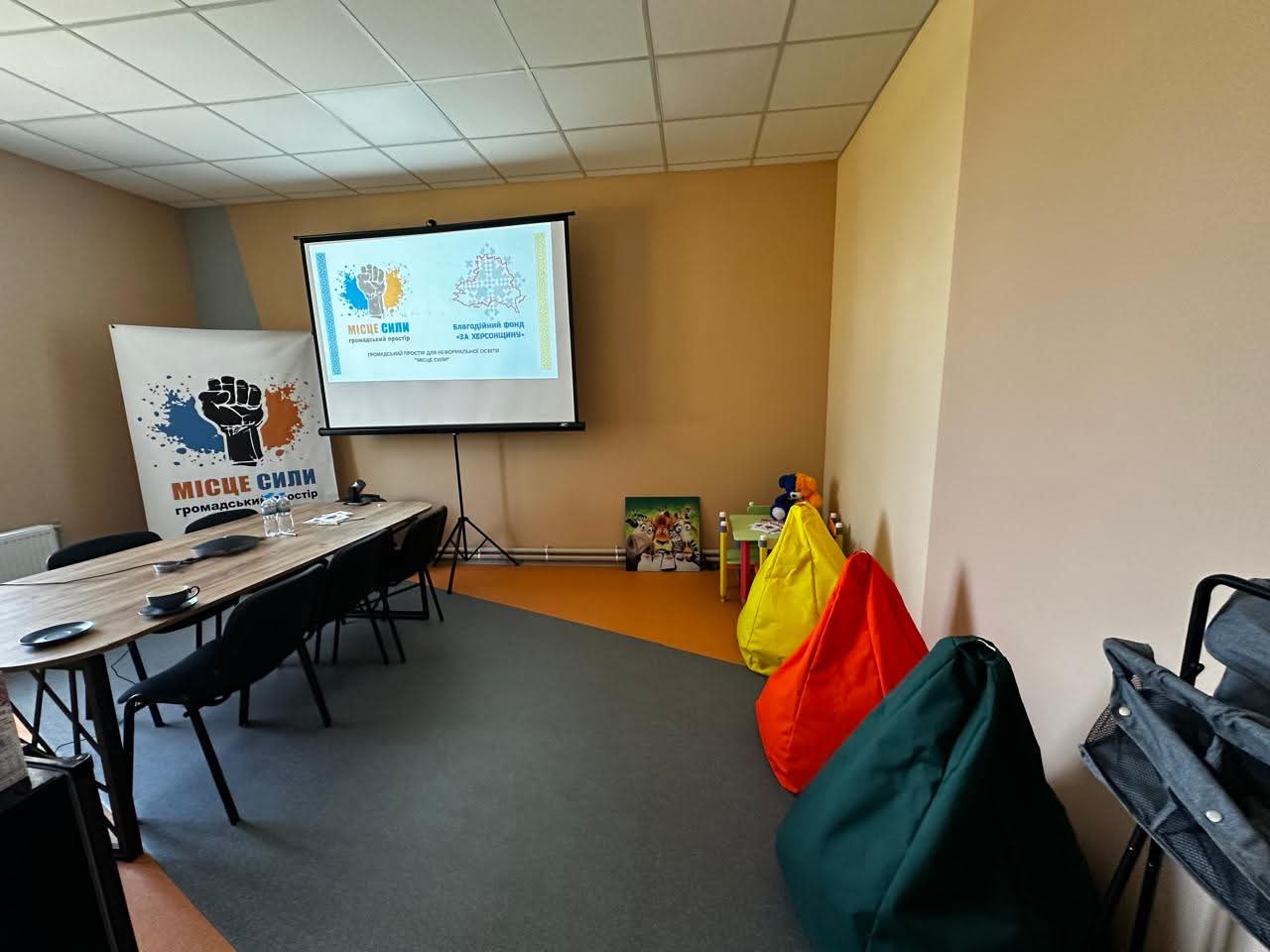 Затишна кімната для зустрічей або навчання з проектором, на екрані якого відображені логотипи: Місце сили та Благодійний фонд На Херсонщині, довгий стіл із кріслами, банер МІСЦЕ СИЛИ ГРОМАДСЬКИЙ ПРОСТІР та три різнокольорові крісла-мішки.