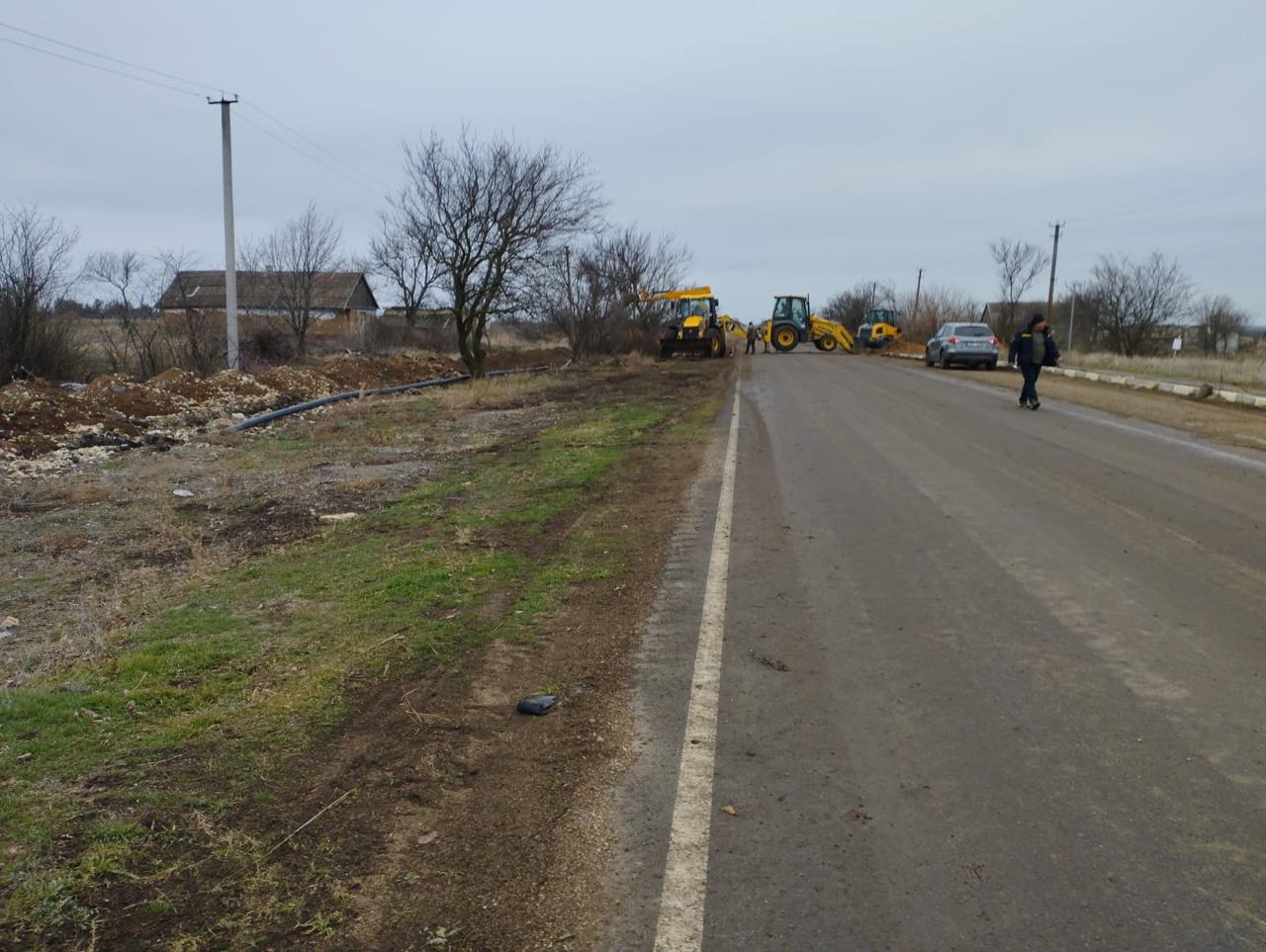 Кадр із ремонту дороги або комунікацій у сільській місцевості. Вздовж узбіччя стоять кілька одиниць будівельної техніки (екскаватори/трактори), а одна людина йде по мокрій асфальтованій дорозі.