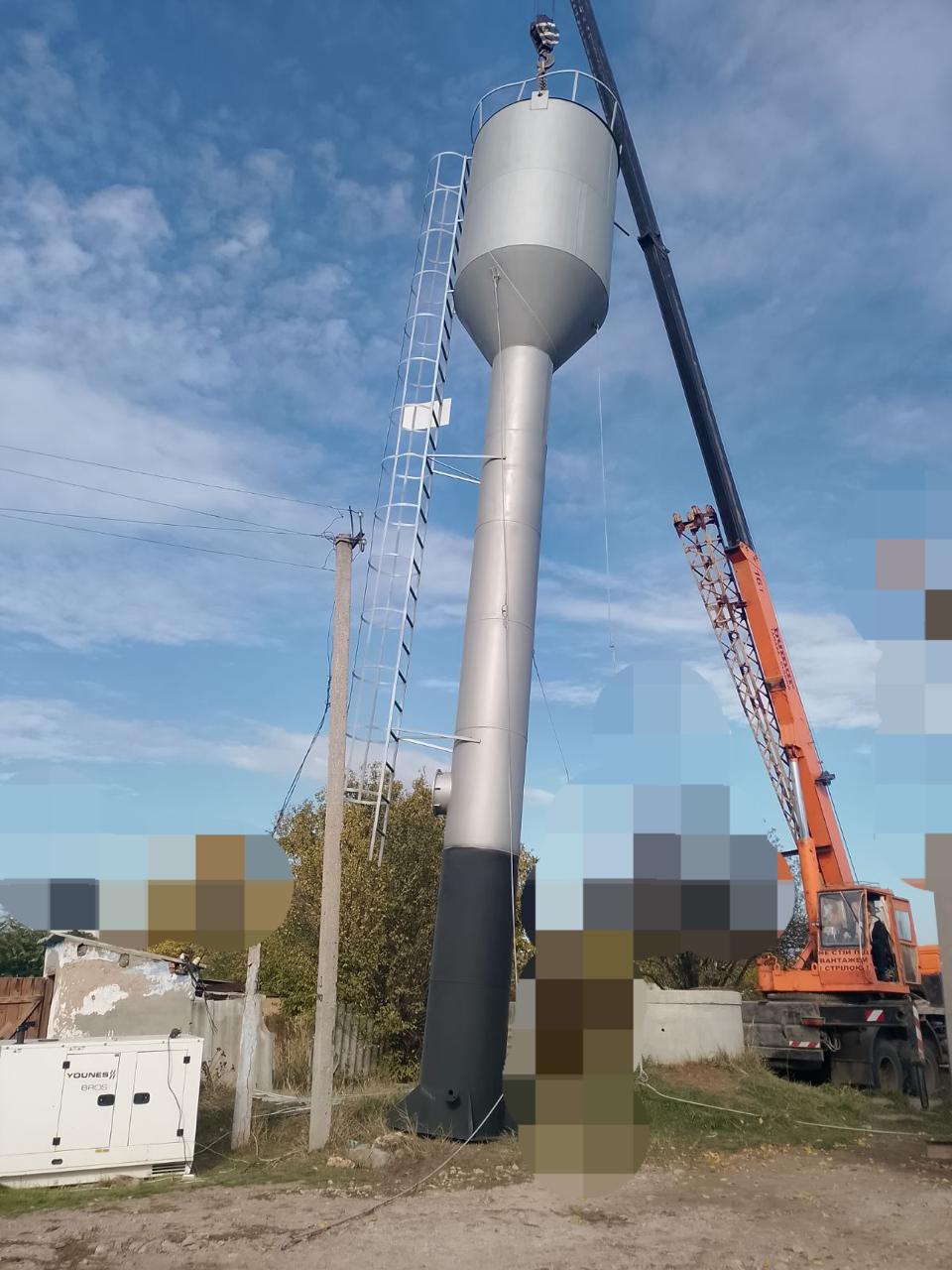 Висока нова металева водонапірна вежа, яку підіймає або встановлює великий помаранчевий автокран під блакитним небом. Поруч стоїть білий дизельний генератор.