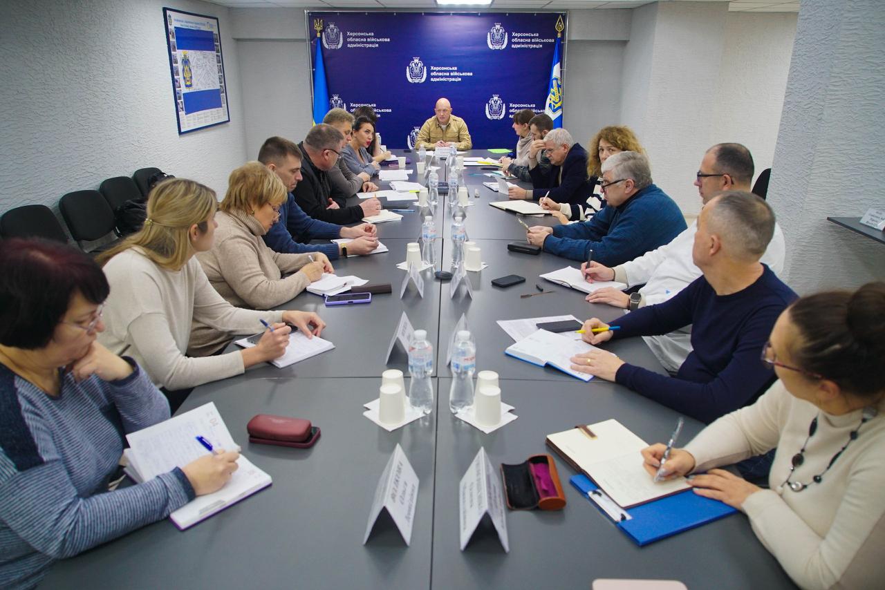 Група людей сидить за довгим прямокутним столом у конференц-залі, проводячи нараду. Більшість учасників зосереджено пишуть. На стіні за центральною особою розташовані таблички з логотипами Херсонської обласної військової адміністрації.
