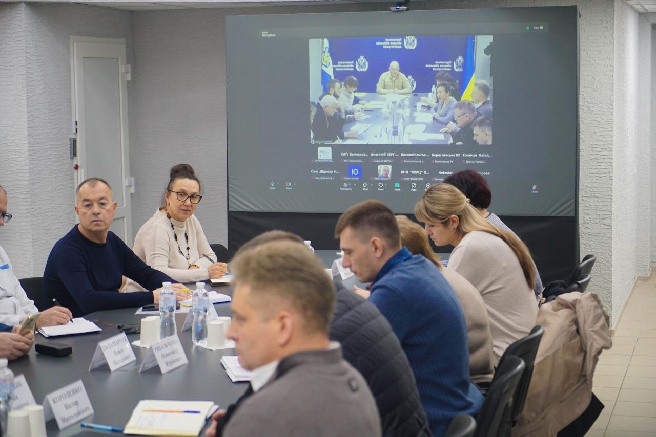 Група людей сидить за столом на нараді, дивлячись на великий екран, де відбувається відеоконференція з іншими учасниками.