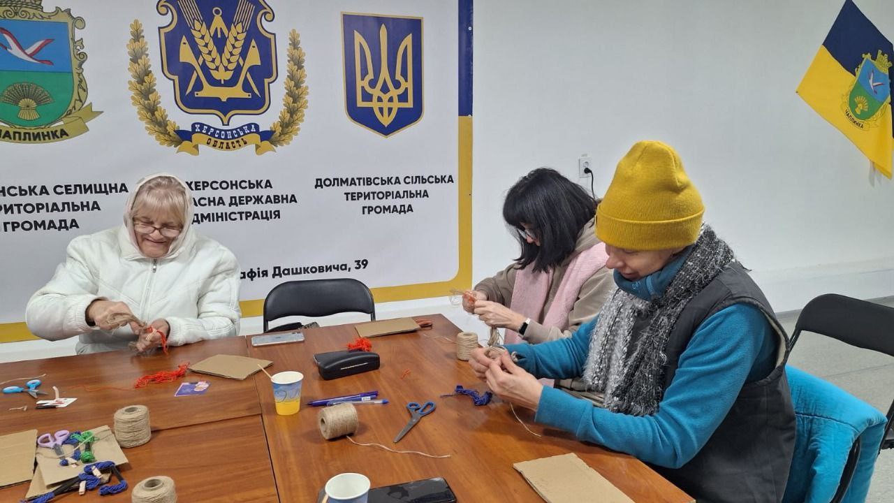 Троє жінок різного віку займаються рукоділлям за дерев'яним столом. Жінки скручують нитки або мотузки, ймовірно, створюючи прикраси чи обереги. На столі видно бобіни зі шпагатом, ножиці та картон. На задньому плані розташований стенд із символікою українських громад: Чаплинської селищної, Херсонської обласної державної адміністрації та Долматівської сільської територіальної громади.