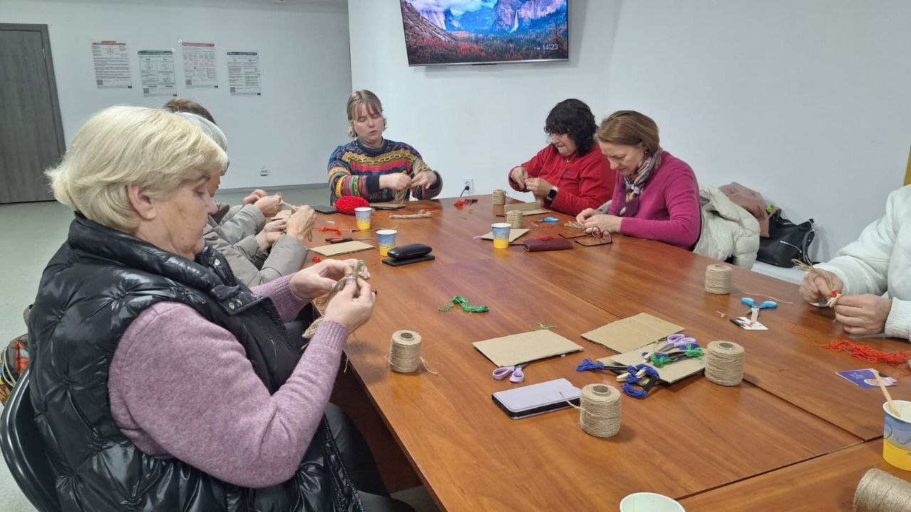 Група жінок різного віку сидить за довгим дерев'яним столом у приміщенні та займається рукоділлям. Жінки скручують нитки або шпагат, виготовляючи ляльки-мотанки або фігурки. На столі лежать ножиці, картон, бобіни зі шпагатом та різнокольорові нитки. На задньому плані розташований великий телевізор, на якому відображається пейзаж.