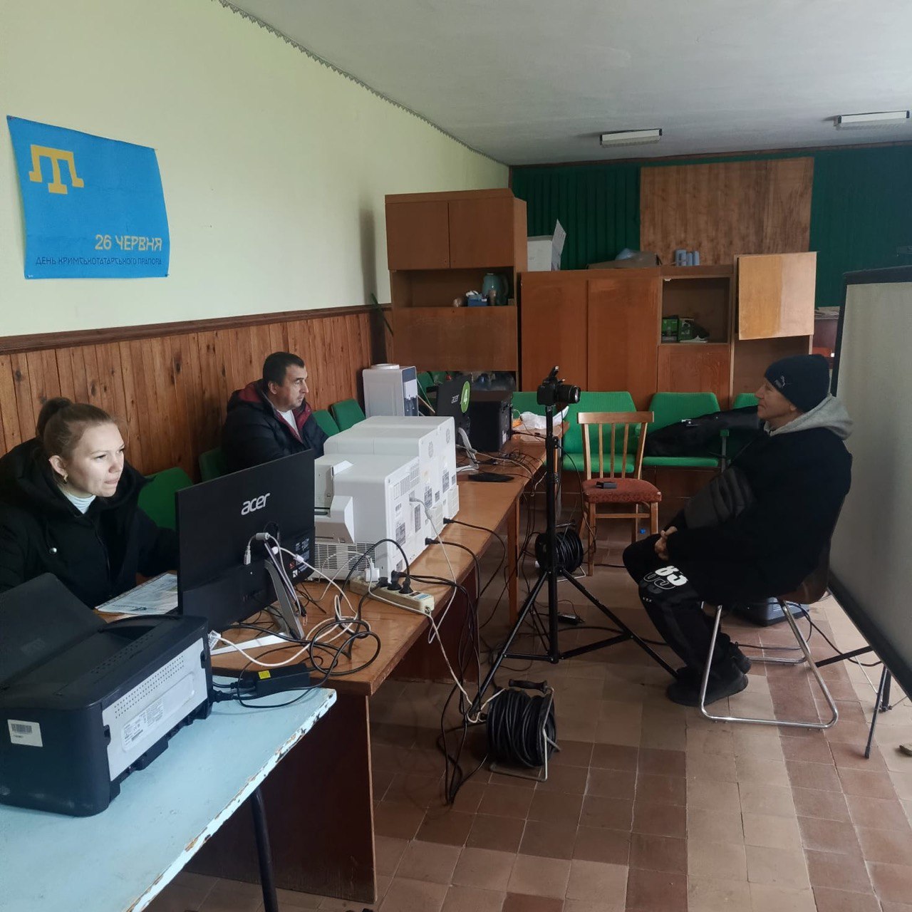 Двоє чоловіків та жінка сидять за столом у приміщенні. Жінка та чоловік ліворуч працюють за комп'ютерами, а чоловік у чорній шапці праворуч сидить навпроти них.