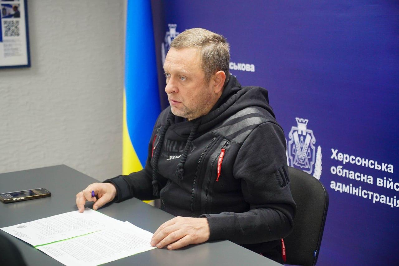 Чоловік у чорній спортивній кофті сидить за столом і розглядає документи. Позаду нього розташовані сині банери з державними прапорами України та символікою Херсонської обласної військової адміністрації.