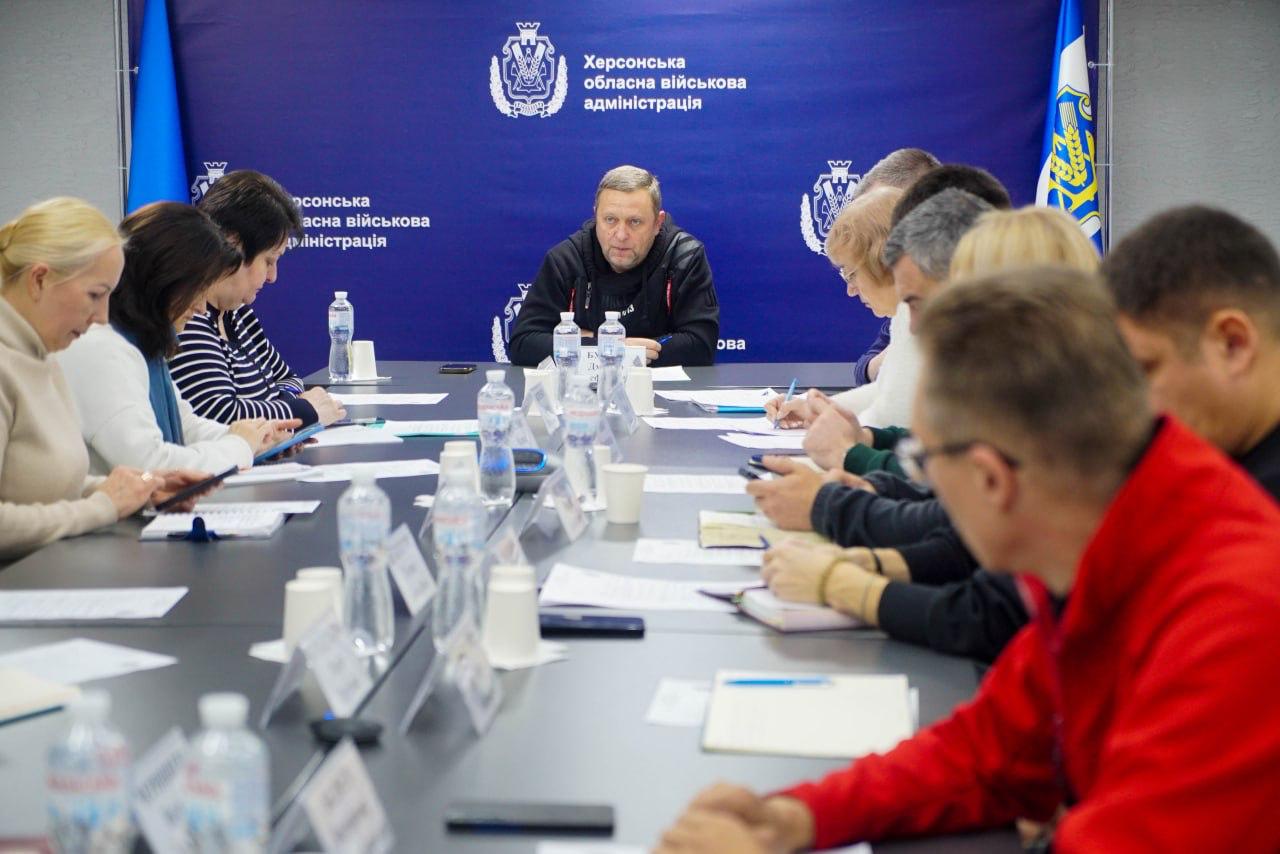 Група людей сидить за великим столом у приміщенні Херсонської обласної військової адміністрації на нараді. Чоловік у центрі сидить обличчям до камери, а інші учасники розташовані з боків, роблячи нотатки.
