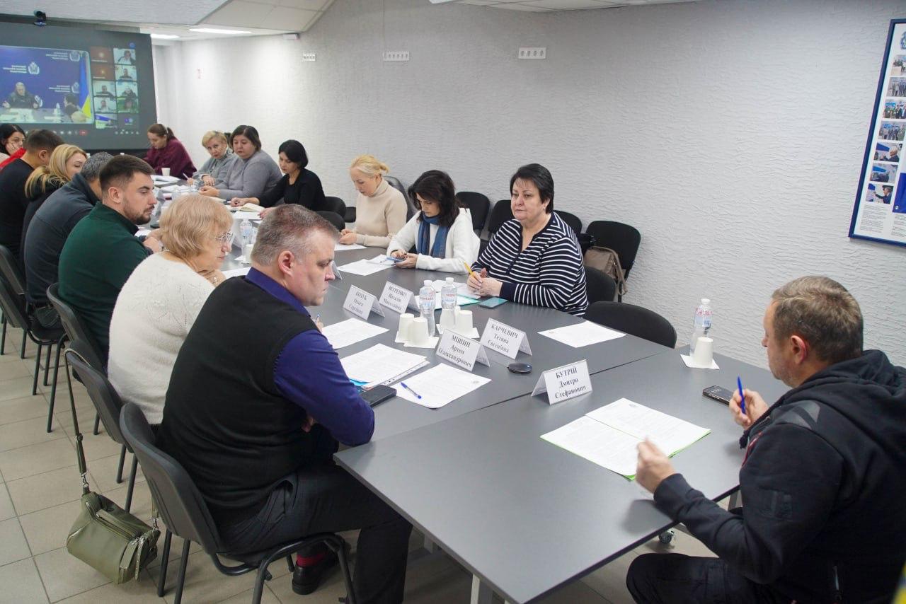 Група людей сидить за великим столом на нараді, обличчям до чоловіка в чорній кофті, який сидить у центрі на тлі синього банера з символікою Херсонської обласної військової адміністрації.
