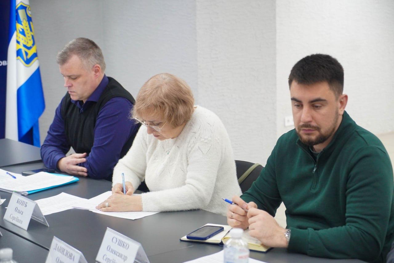 Троє людей — чоловік у темно-синьому светрі, жінка у білому светрі та чоловік у темно-зеленому светрі — сидять за столом на нараді, пишуть або переглядають документи. Усі троє розташовані на тлі прапорів та синього банера.