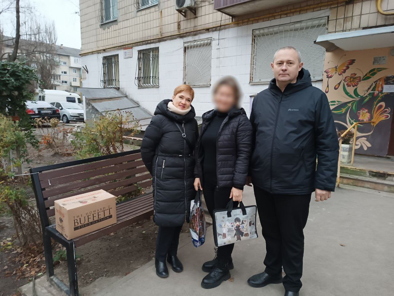 Дві жінки та чоловік стоять на вулиці перед багатоповерховим будинком. На лавці поруч стоїть коричнева коробка з логотипом The Howard G. Buffett Foundation.