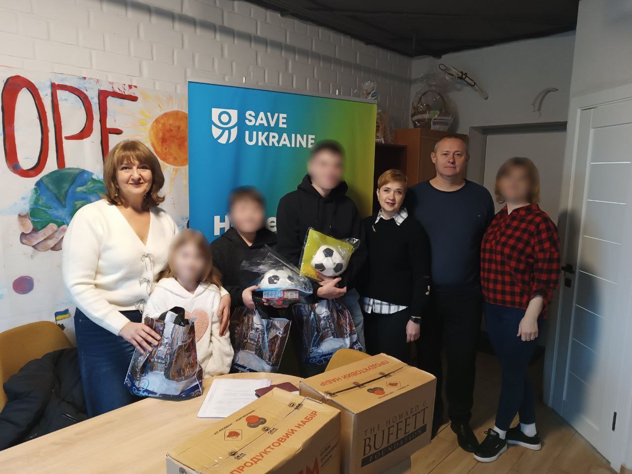 Групова фотографія, де дорослі та діти стоять біля банера SAVE UKRAINE. На передньому плані видно коробки з написами: ПРОДУКТОВИЙ НАБІР та THE HOWARD G. BUFFETT FOUNDATION. Двоє хлопчиків тримають у руках футбольні м'ячі.